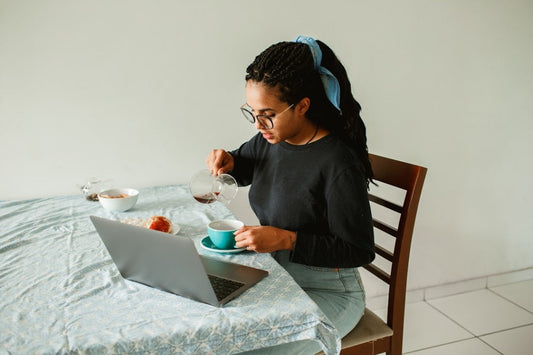 Young woman working on a laptop while pouring tea at the kitchen table, showing a simple way to balance wellness with a busy lifestyle.