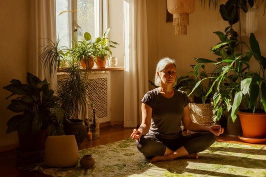 Older woman meditating cross-legged in a sunlit room filled with houseplants.