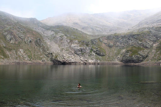 Person swimming alone in a tranquil mountain lake surrounded by rocky cliffs and mist.