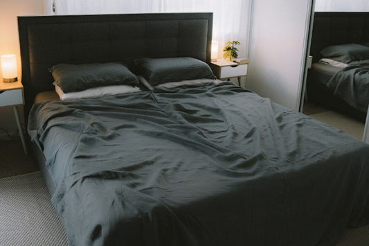 A neatly made bed with dark grey sheets in a softly lit, minimalist bedroom.