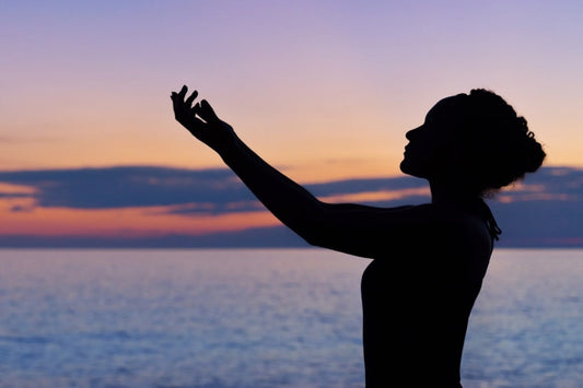 Silhouette of a woman raising her hands toward the sky at sunset by the ocean.