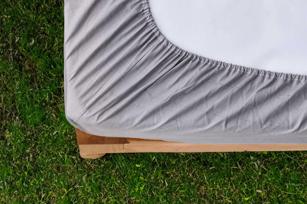 Close-up of a gray fitted grounding sheet corner on a wooden bed frame set over green grass.