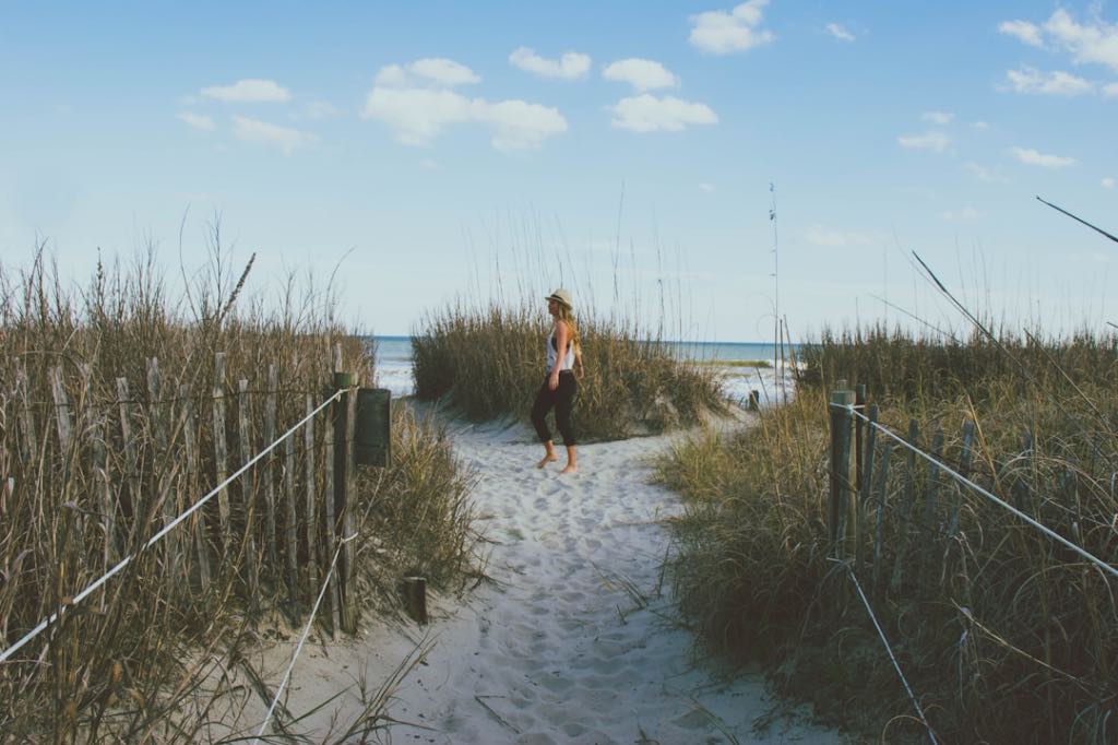 Woman walking barefoot on a sandy path through beach grass, heading toward the ocean.