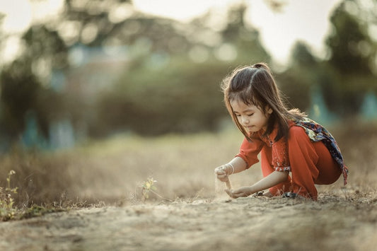 Young girl in a red outfit crouching outdoors, playing with soil in a natural setting.