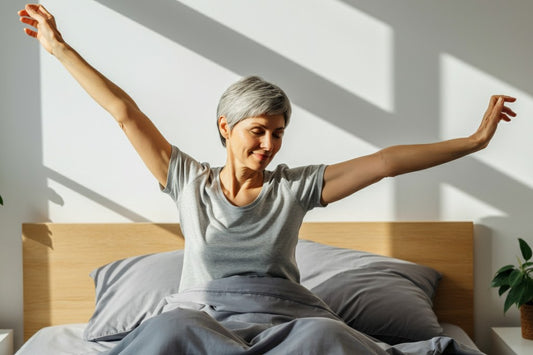 Woman with short gray hair stretching happily in bed after waking up, sunlight streaming through the window.