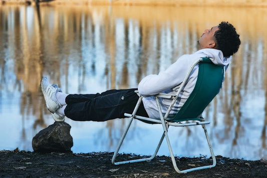 Man relaxing by a lake in a green folding chair with feet propped on a rock, eyes closed in stillness.