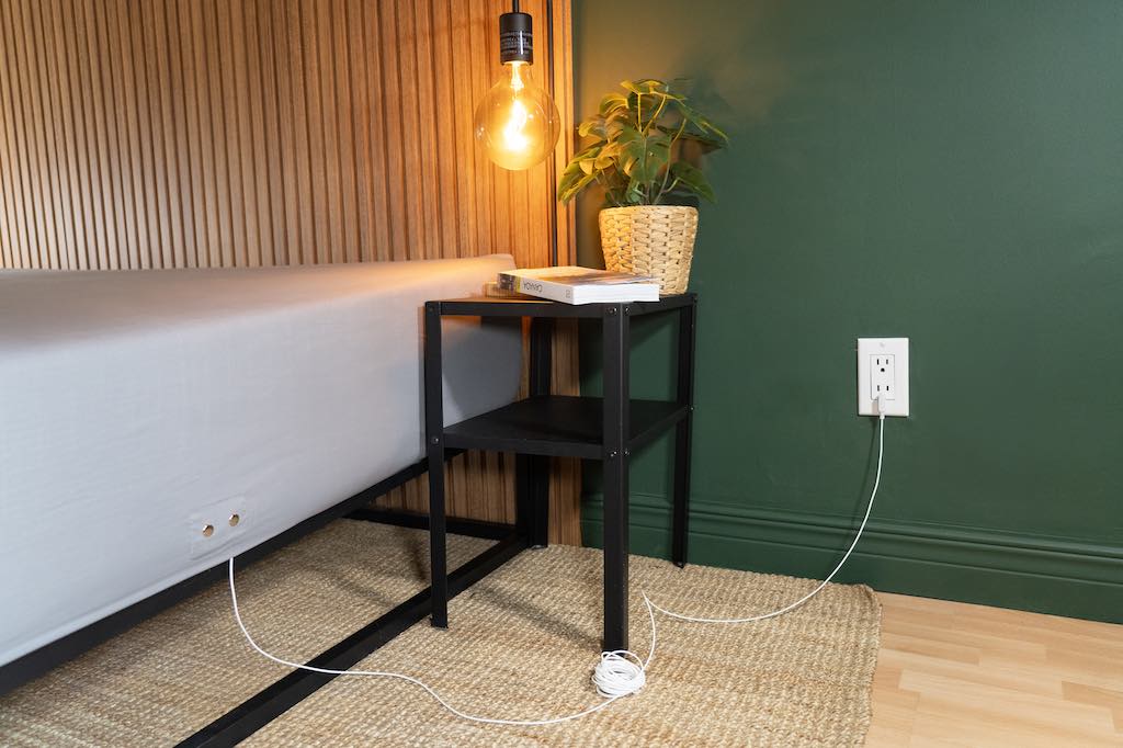 Grounding sheet plugged into a wall outlet beside a bed with a small black nightstand and warm lighting.