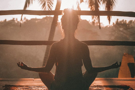 Silhouette of a woman meditating on a wooden deck at sunrise, surrounded by palm trees.