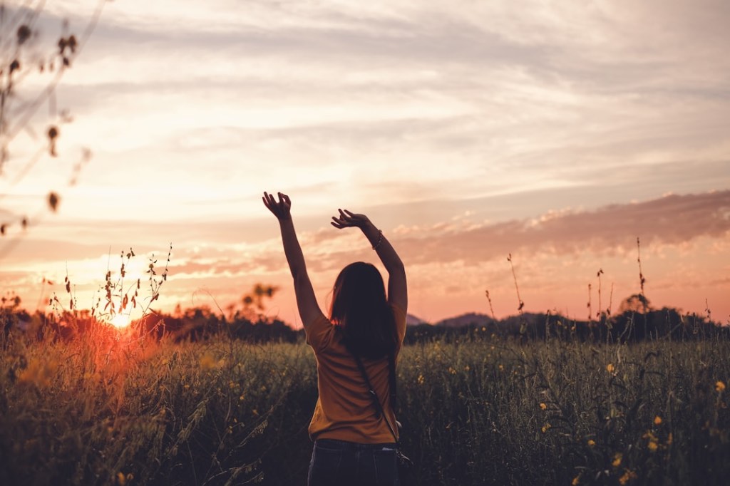 Woman standing in a wildflower field at sunset with arms raised toward the sky.