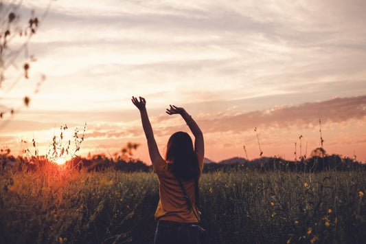 Woman standing in a wildflower field at sunset with arms raised toward the sky.