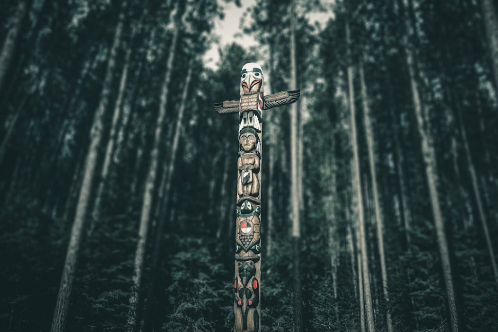 Tall, painted Indigenous totem pole standing in a dense, dark green forest.